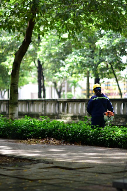 A gardener in protective clothing, including a helmet and dark uniform, is seen trimming a well-maintained hedge along a paved sidewalk in a park or garden area. The scene is shaded by large, lush gre