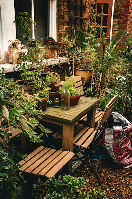 A cozy urban garden courtyard featuring a weathered wooden dining table surrounded by matching wooden and metal chairs, set against a brick building with a white-framed window. The table displays vari