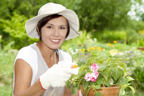 A woman with light skin, wearing a wide-brimmed white gardening hat, white gardening gloves, and a light-colored top, is smiling while carefully tending to a pot of pink and white flowering plants in a lush outdoor garden. The garden environment features a well-maintained lawn with dense, green grass in the foreground, and various shrubs and flowering plants in the background, creating a vibrant and natural setting. The scene is bathed in natural daylight, indicating a bright, possibly partly cloudy outdoor day, with out-of-focus greenery and colorful blooms highlighting the active gardening work. This image exemplifies typical gardening practices suitable for residential outdoor spaces in East London, such as those serviced by Gardener Newham, focusing on planting and garden maintenance activities in a landscaped backyard or front garden environment.